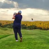 En golfare svingar klubban på en utslagsplats i golf. Solstrålar bryter igenom molnen över en grönskande bana.