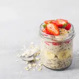En burk med overnight oats recept toppad med jordgubbar och banan. En sked ligger bredvid med havregryn.