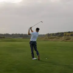 En man svingar sin golfklubba på en grön golfbana. Frågan "hur lång tid tar en golfrunda" kan besvaras med att det beror på många faktorer.