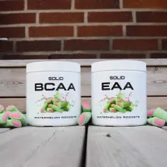 Två burkar, en med BCAA och en med EAA, står framför en tegelvägg. Gröna godisar med rosa inre ligger utspridda.