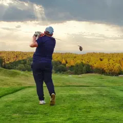 En golfare svingar klubban på en utslagsplats i golf. Solstrålar bryter igenom molnen över en grönskande bana.