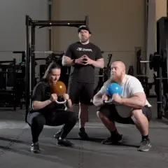 Två personer utför goblet squats med kettlebells, en instruktör står bakom.