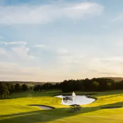 Solnedgång över en golfbana med en damm och fontän. En spelare är på väg att slå ett slag, kanske för en albatross.