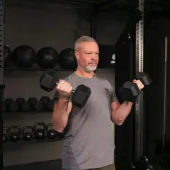 Man med grått hår och skägg utför sittande bicepscurl med hantlar i ett gym.