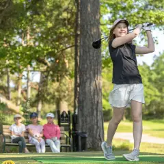 En flicka svingar sin golfklubba på ett soligt golfhål. Tre barn sitter på en bänk i bakgrunden och tittar på.
