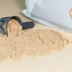 Mörkblå skopa med ljusbrunt pulver, en del av det har spillts ut. Påsen i bakgrunden har en brun etikett med texten "MOCHA". Detta visar vad som kan finnas i proteinpulver.