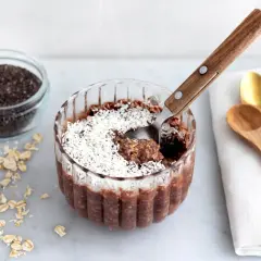 En skål med overnight oats chokladboll, toppad med kokosflingor. Bredvid finns ingredienser som havregryn, chiafrön och kakaopulver.