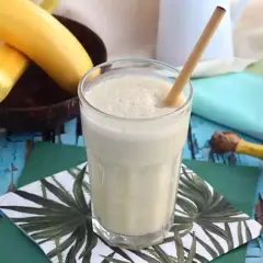 En krämig banan milkshake med en bambustrå i ett glas, omgiven av bananer och en kokosnöt.