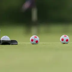 En golfputter är redo att slå en boll med fotbollsmönster. Två likadana bollar ligger längre bort på greenen.