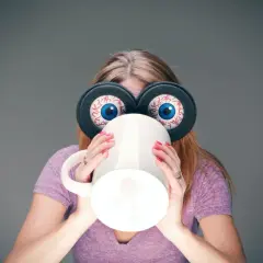 Kvinna med stora, uppspärrade ögon och en mugg framför ansiktet, en bild av koffeinberoende.