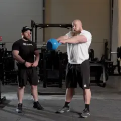 Man med skägg utför kettlebell swing varje dag i gymmet. Bredvid står en tränare.