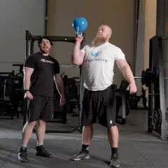 Man med skägg utför en kettlebell press med en blå kettlebell. En annan man står bredvid och tittar.