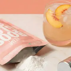 En påse "Glow Boost" med persikosmak, en drink med persika, och en hantel. Frågan "hur mycket kreatin per dag" kan besvaras med denna produkt.