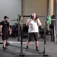 Man med skägg förbereder sig för knäböj med en safety squat bar. En tränare står bredvid.
