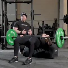 Man instruerar kvinna som utför hip thrust med skivstång på en bänk i gym.