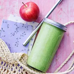 En grön, proteinrik smoothie i glasburk med metallrör, bredvid ett rött äpple och en vävd kasse på en rosa bakgrund.