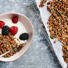 En skål med yoghurt, bär och hemgjord granola – en perfekt frukost utan kolhydrater. Granola med nötter och frön ligger bredvid på ett bakplåtspapper.