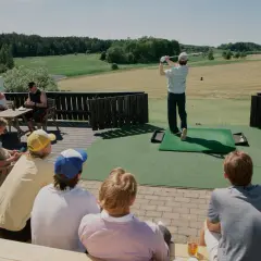 En solig dag på golfbanan. En person slår ett slag medan andra sitter och tittar på, kanske diskuterar annorlunda spelformer golf.