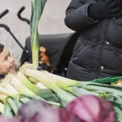 Glad pojke tittar på purjolök och lök på en marknad. En person i svart jacka håller i en grön låda med billiga råvaror.