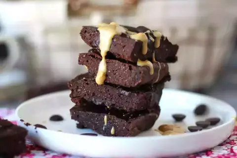 En stapel mörka protein brownies med en krämig topping och chokladknappar på en vit tallrik.