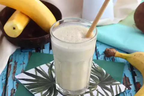 En krämig banan milkshake med en bambustrå i ett glas, omgiven av bananer och en kokosnöt.