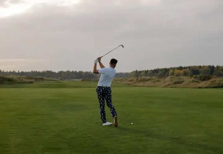 En man svingar sin golfklubba på en grön golfbana. Frågan "hur lång tid tar en golfrunda" kan besvaras med att det beror på många faktorer.