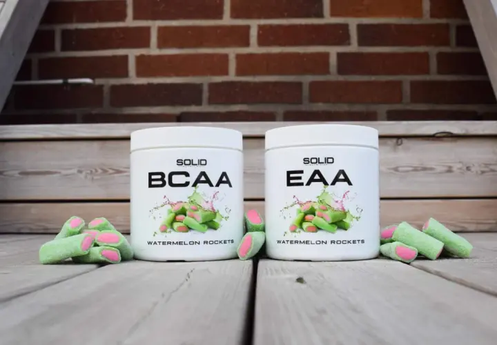 Två burkar, en med BCAA och en med EAA, står framför en tegelvägg. Gröna godisar med rosa inre ligger utspridda.