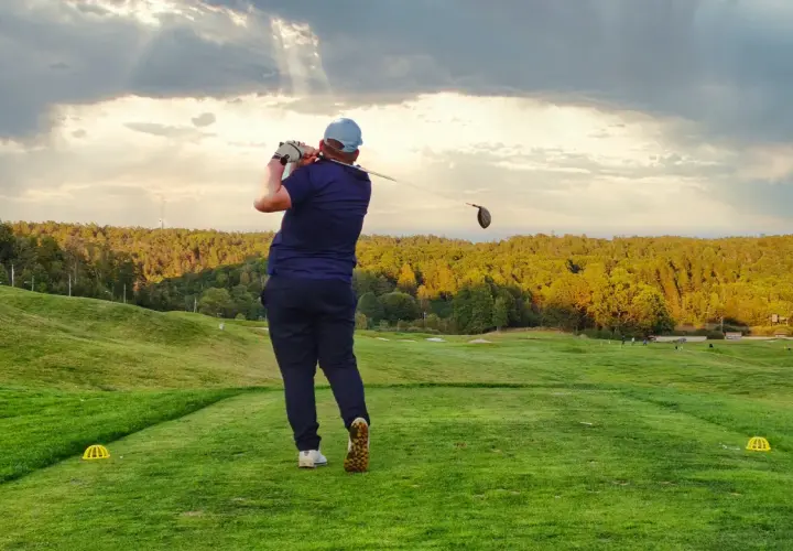 En golfare svingar klubban på en utslagsplats i golf. Solstrålar bryter igenom molnen över en grönskande bana.