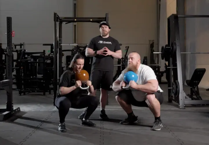 Två personer utför goblet squats med kettlebells, en instruktör står bakom.