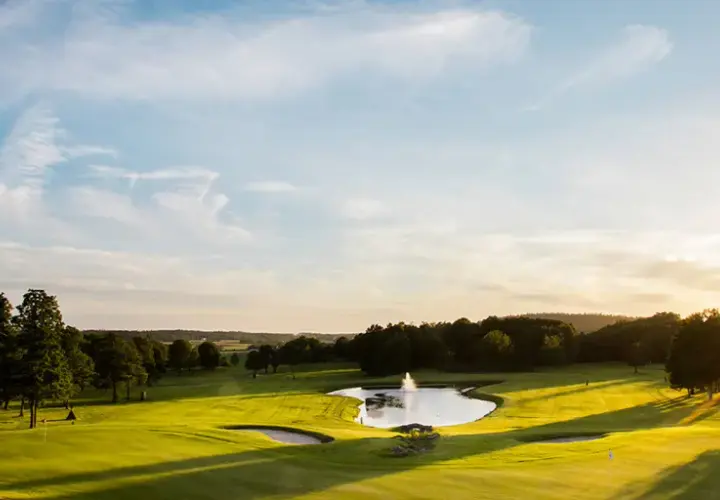 Solnedgång över en golfbana med en damm och fontän. En spelare är på väg att slå ett slag, kanske för en albatross.