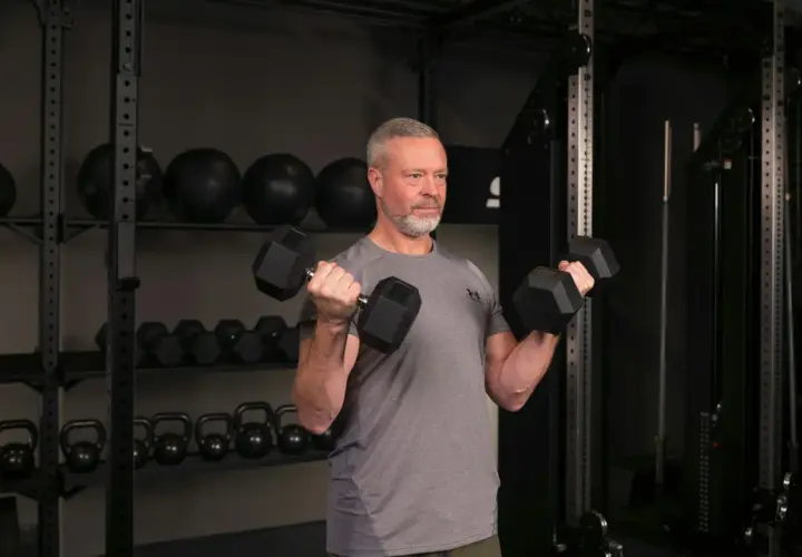 Man med grått hår och skägg utför sittande bicepscurl med hantlar i ett gym.