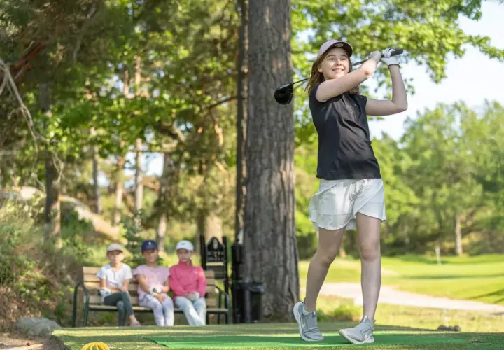 En flicka svingar sin golfklubba på ett soligt golfhål. Tre barn sitter på en bänk i bakgrunden och tittar på.