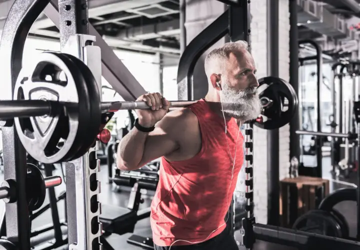 Äldre man med vitt skägg tränar med skivstång. Han lyssnar på musik. Träning motverkar sarkopeni, vilket betyder muskelförlust.