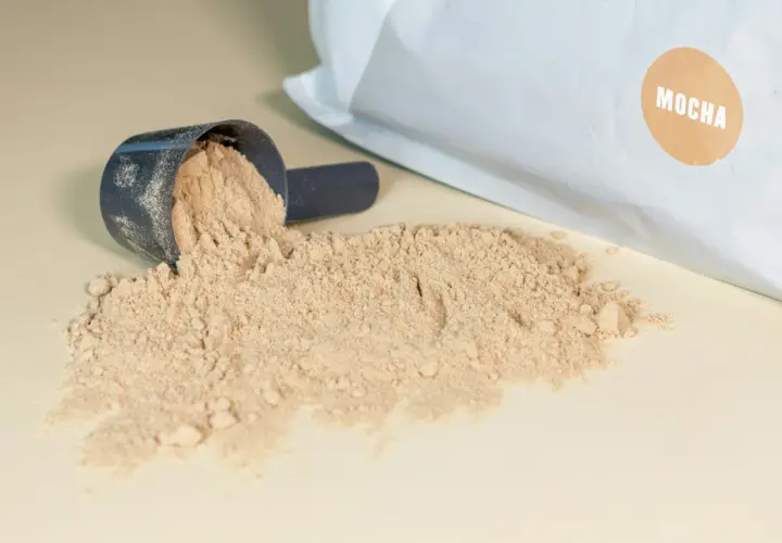 Mörkblå skopa med ljusbrunt pulver, en del av det har spillts ut. Påsen i bakgrunden har en brun etikett med texten "MOCHA". Detta visar vad som kan finnas i proteinpulver.