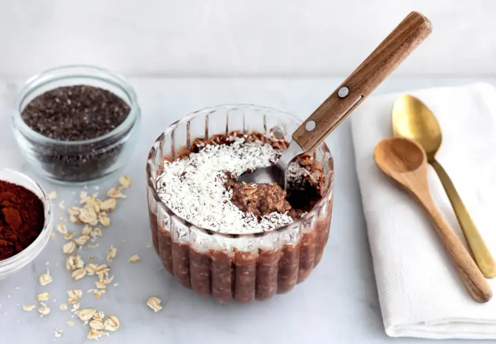 En skål med overnight oats chokladboll, toppad med kokosflingor. Bredvid finns ingredienser som havregryn, chiafrön och kakaopulver.
