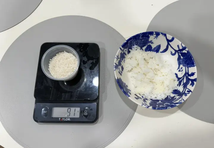 En digitalvåg visar 91 gram ris i en liten behållare. Bredvid står en skål med mer ris, redo att ätas. Perfekt för att hålla koll på råris kalorier.