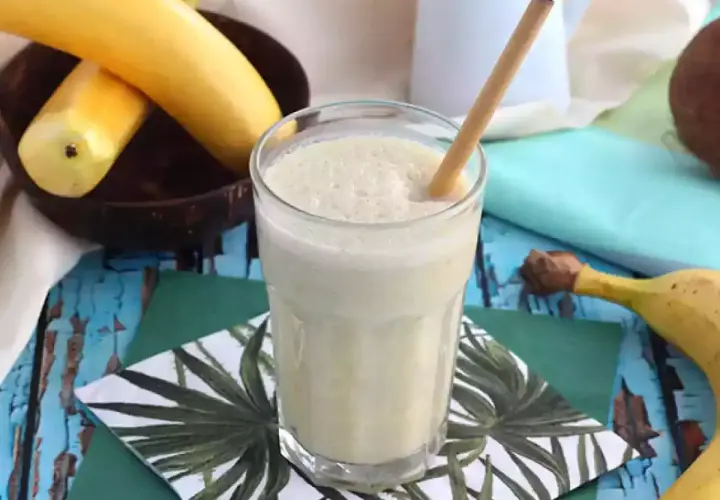 En krämig banan milkshake med en bambustrå i ett glas, omgiven av bananer och en kokosnöt.