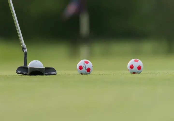 En golfputter är redo att slå en boll med fotbollsmönster. Två likadana bollar ligger längre bort på greenen.