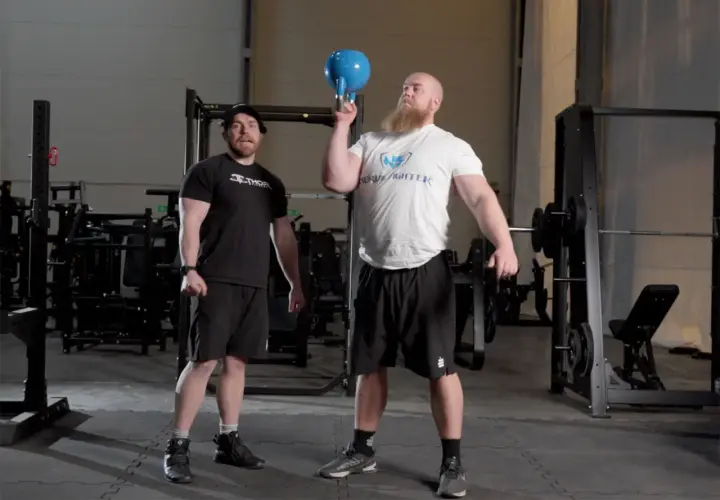 Man med skägg utför en kettlebell press med en blå kettlebell. En annan man står bredvid och tittar.