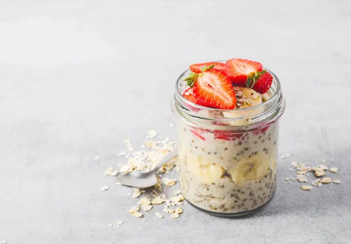 En burk med overnight oats recept toppad med jordgubbar och banan. En sked ligger bredvid med havregryn.