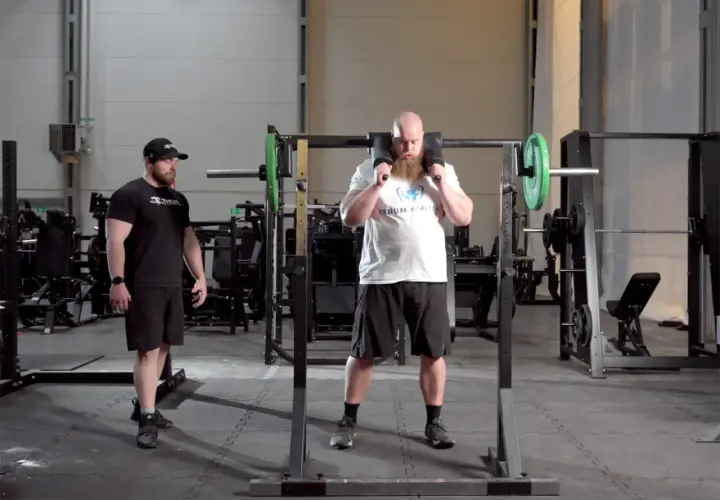 Man med skägg förbereder sig för knäböj med en safety squat bar. En tränare står bredvid.