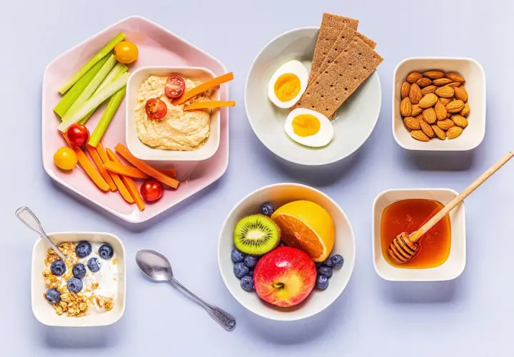 En frukost med kolhydratrik mat: yoghurt med granola och blåbär, frukt, ägg, kex, nötter, honung och grönsaksstavar med hummus.