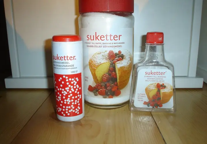 Tre förpackningar Suketter, ett sockerersättningsmedel, står på ett träbord. En burk, en tub och en flaska. Suketter är utmärkt till bakning och matlagning, helt utan kalorier.