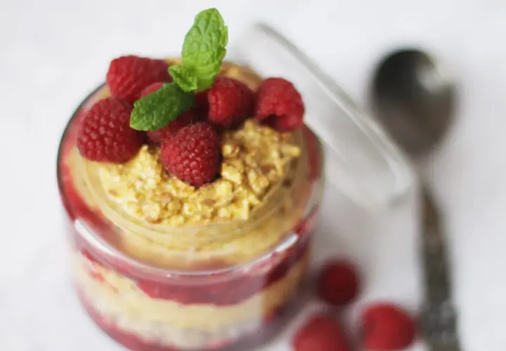 En burk med overnight oats med saffranssmak, toppad med hallon och mynta. En sked ligger bredvid.