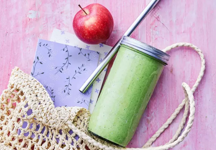 En grön, proteinrik smoothie i glasburk med metallrör, bredvid ett rött äpple och en vävd kasse på en rosa bakgrund.