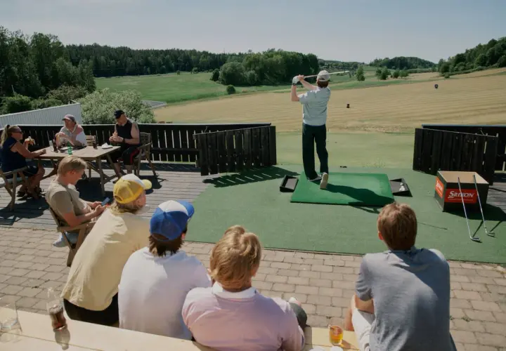En solig dag på golfbanan. En person slår ett slag medan andra sitter och tittar på, kanske diskuterar annorlunda spelformer golf.