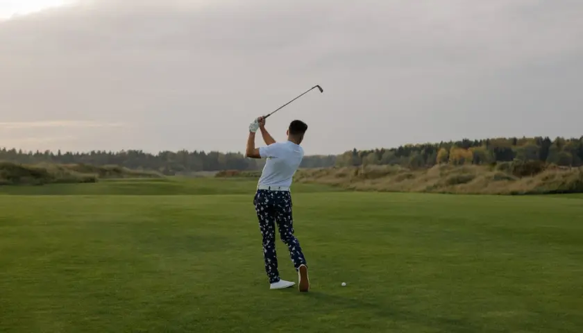 En man svingar sin golfklubba på en grön golfbana. Frågan "hur lång tid tar en golfrunda" kan besvaras med att det beror på många faktorer.