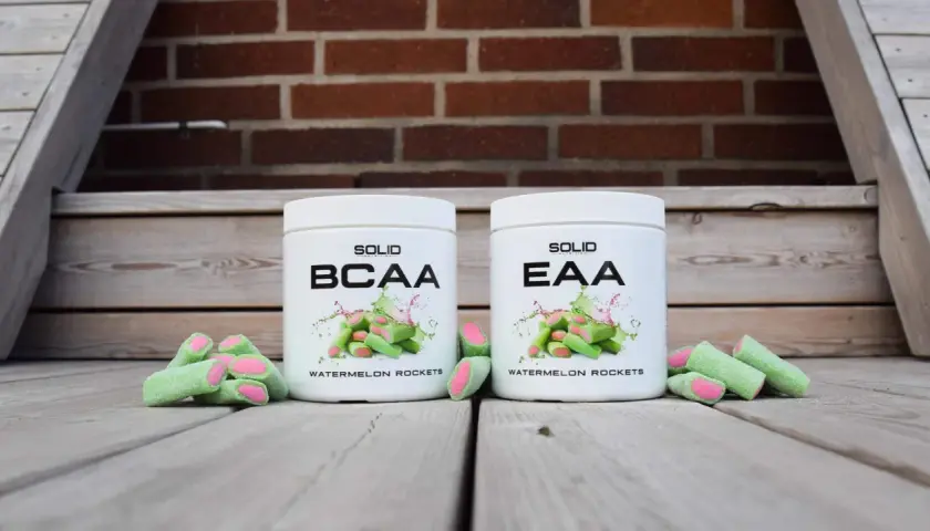Två burkar, en med BCAA och en med EAA, står framför en tegelvägg. Gröna godisar med rosa inre ligger utspridda.