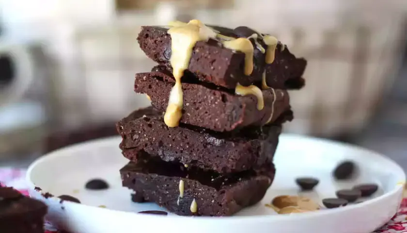 En stapel mörka protein brownies med en krämig topping och chokladknappar på en vit tallrik.