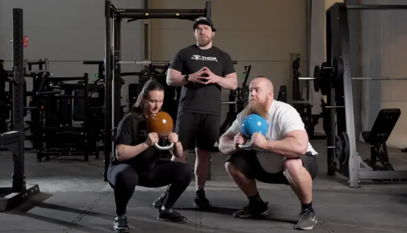 Två personer utför goblet squats med kettlebells, en instruktör står bakom.